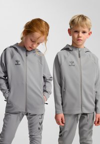 Grå atletiske hættetrøjer med lynlås og logo, kombineret med matchende bukser. Begge har en glat tekstur og en tilspidset pasform, designet til børn.