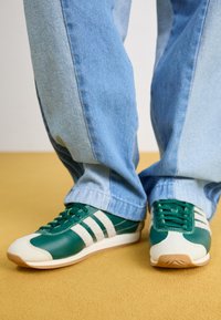 Zielone skórzane sneakersy z białymi paskami, zaokrąglonym noskiem i teksturowanymi akcentami. Połączone z jasnoniebieskimi dżinsami. Podłoże jest żółte.