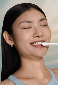 Brosse à dents électrique avec un manche blanc et des poils bleus tenue près d'une bouche souriante avec des dents saines et blanches, sur un fond doux.