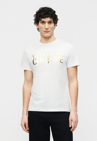 Νέος άνδρας με σγουρά μαλλιά, φοράει λευκό t-shirt Versace Jeans Couture με χρυσά γράμματα και μαύρο παντελόνι, στέκεται μπροστά σε απλό φόντο.
