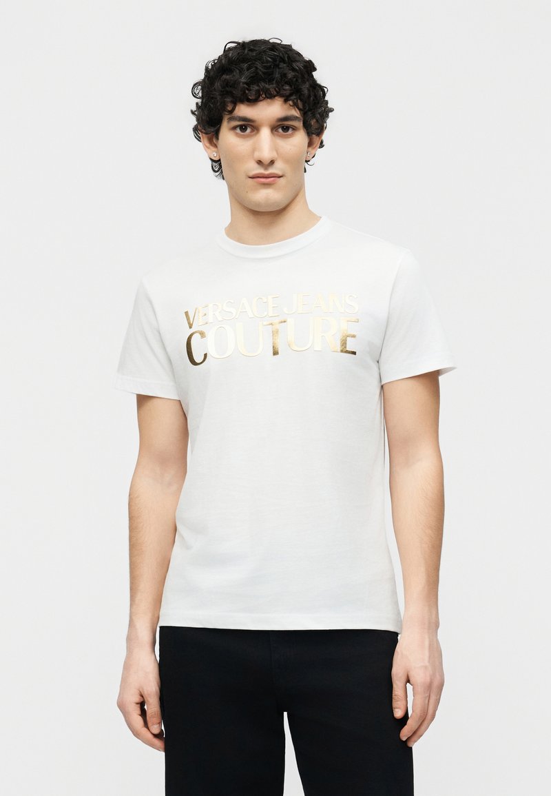 Νέος άνδρας με σγουρά μαλλιά, φοράει λευκό t-shirt Versace Jeans Couture με χρυσά γράμματα και μαύρο παντελόνι, στέκεται μπροστά σε απλό φόντο.