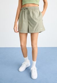 Zelené Nike šortky s elastickým pásom a šnúrkou, obsahujúce bočné vrecká a malé logo na okraji. Vytvorené v kombinácii s bielymi teniskami.