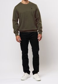 Oliivinvihreä pyöreäkaulusneule, jossa on pitkät hihat, pieni musta logo ja yhdistetty mustiin slim-fit-housuihin sekä valkoisiin lenkkarit.