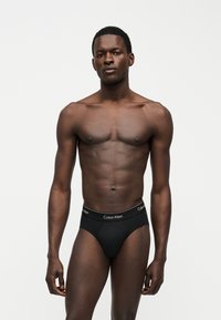 Mannequin masculin à la peau foncée portant un slip noir Calvin Klein, debout devant un fond blanc uni, regardant droit avec une expression neutre.