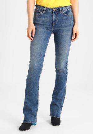 Femme portant un jean bootcut bleu et des chaussures noires debout sur un fond blanc.