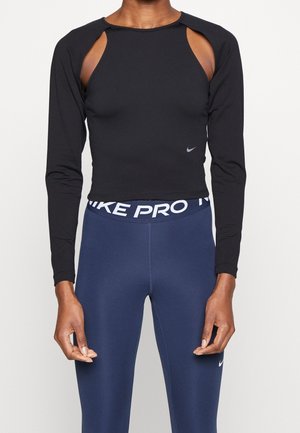 Personne portant un crop top noir à manches longues avec découpes aux épaules et des leggings Nike Pro bleu marine avec une ceinture visible avec le logo.