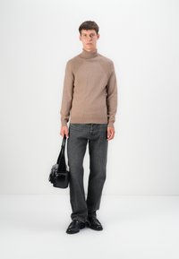 Pull à col roulé brun en maille avec détails côtelés, associé à un jean gris foncé et des chaussures en cuir noir. Le mannequin tient un sac bandoulière noir.