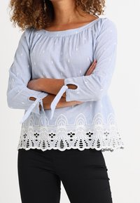 Haut à manches longues bleu clair avec des accents brodés blancs et une finition en dentelle au bas. Présente des liens aux manches et une encolure dégagée.