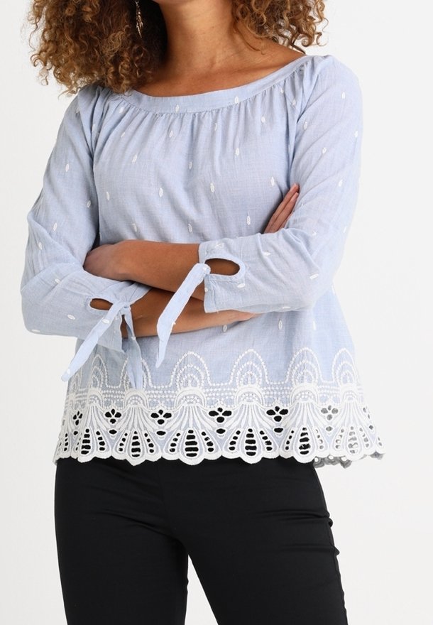 Haut à manches longues bleu clair avec des accents brodés blancs et une finition en dentelle au bas. Présente des liens aux manches et une encolure dégagée.