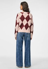 Adīts džemperis ar bordo un rozā argyle rakstu, apaļu kakla izgriezumu un ribotas aproces, saskaņots ar platām zilu džinsu biksēm.