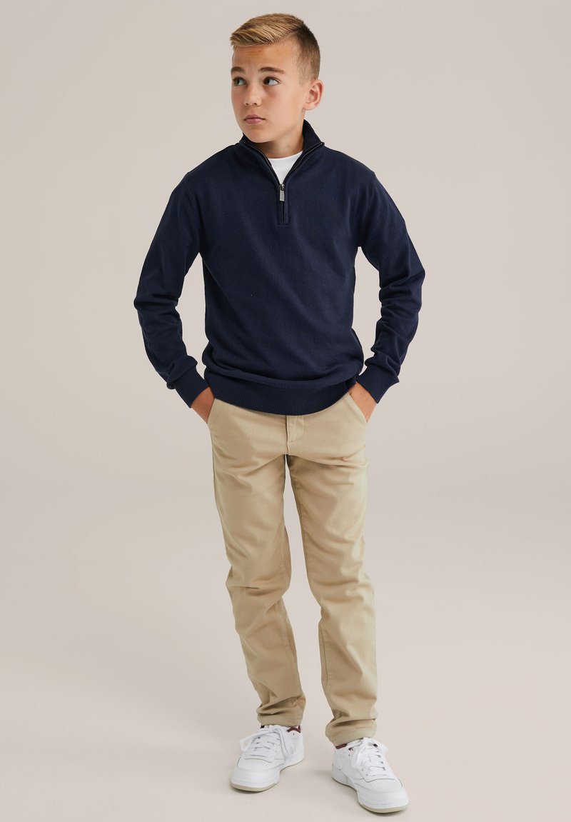 Marinblå zip-up tröja med ribbade muddar, matchad med khakifärgad byxor och vita sneakers. Tröjan har en ståkrage och slim fit.