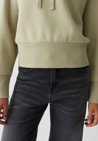 Persona con felpa beige e jeans neri, mani rilassate ai lati, tagliata in vita e a metà coscia.