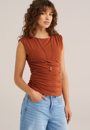 Mujer con cabello rizado que lleva una camiseta sin mangas de color óxido con fruncidos laterales, jeans azul claro y un collar largo con un colgante.