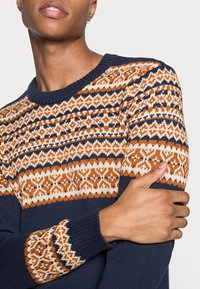 Pull en maille bleu marine avec un empiècement à motifs orange, crème et bleu marine, ornés de dessins complexes sur les manches et les poignets.