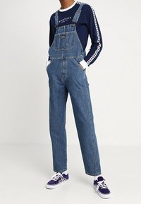 Peto de mezclilla con tirantes ajustables, gran bolsillo frontal y corte holgado. Combinado con una camiseta de manga larga azul marino y zapatillas púrpuras.