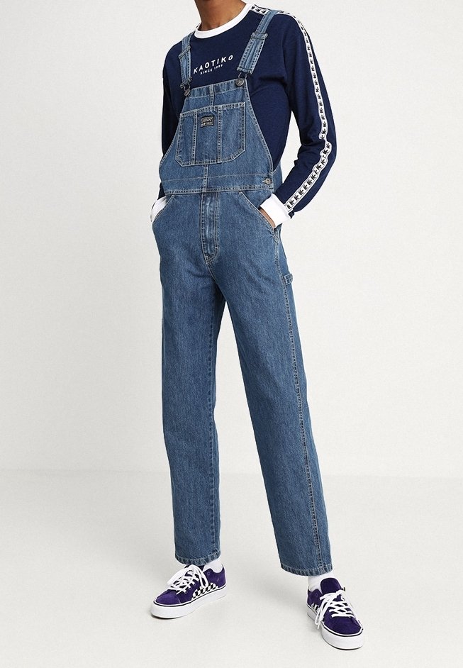 Peto de mezclilla con tirantes ajustables, gran bolsillo frontal y corte holgado. Combinado con una camiseta de manga larga azul marino y zapatillas púrpuras.