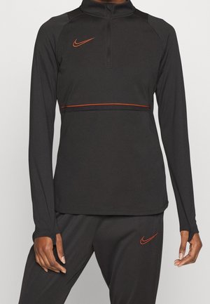 Schwarzes langärmliges Sportoberteil und Hose mit orangen Nike-Swoosh-Logos und einem orangen horizontalen Streifen über der Brust, getragen von einer Person.