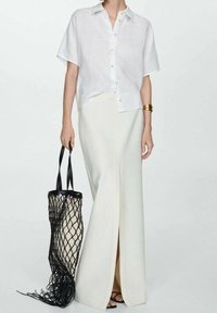 Biała lniana koszula z krótkim rękawem i guzikami, zestawiona z kremową spódnicą do ziemi z rozcięciem z przodu oraz czarną siateczkową torbą typu tote.