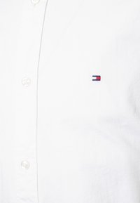 Weißes Baumwoll-Hemd mit Knopfleiste, das ein kleines besticktes Logo in Marineblau und Rot auf der linken Brust zeigt, mit Standardknöpfen.