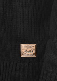 Schwarzer Strickpullover mit geripptem Saum und einem beigefarbenen Lederetikett mit Text. Das Design ist strukturiert und hat eine einheitliche Farbe.