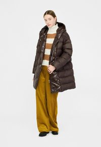 Brun puffy jakke med en shiny tekstur, hætte og knapper, iført over en cremet og brun stribet sweater, parret med gule vide bukser.