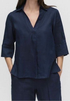 Kobieta w granatowej bluzce z dekoltem w serek, z rękawami do łokci, oraz w dopasowanych granatowych spodniach, z rękami w kieszeniach.