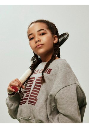 Grå hettegenser med rødt grafisk motiv, flettet hår, holder en tennisracket med hvitt grep og røde detaljer mot en ensfarget bakgrunn.