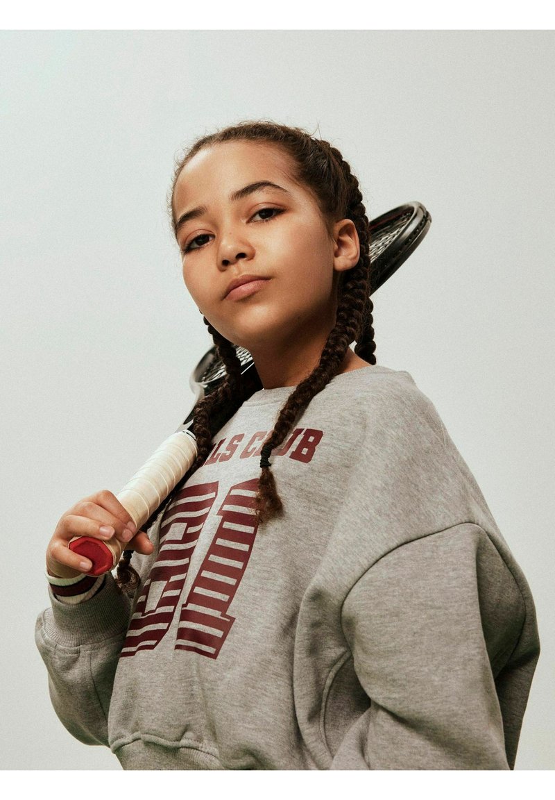 Szara bluza z czerwonym nadrukiem, splecione włosy, trzymająca rakietę tenisową z białym uchwytem i czerwonymi akcentami na jednolitym tle.