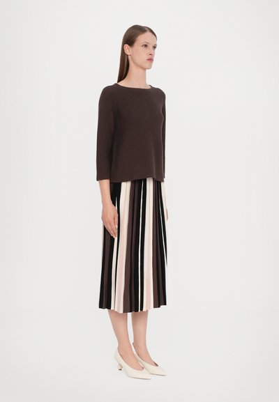 Pull en maille marron à manches trois quarts, associé à une jupe plissée rayée dans des nuances de noir, beige et rose ; talons aiguilles blancs.
