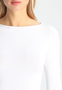 White long-sleeve top with a round neckline, featuring a smooth, stretchy fabric and minimalistic design; appears form-fitting.