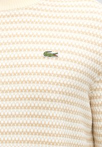 Tissu tricoté beige et blanc texturé avec un petit logo crocodile vert brodé sur la partie supérieure de la poitrine.