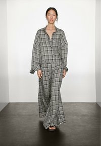 Modelka sebevědomě kráčí v volném kostkovaném šedém outfitu, který zahrnuje košilku na knoflíky a široké kalhoty, na jednoduchém pozadí.