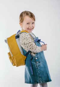Zaino giallo con spallacci blu, fissato di lato. Bambina che indossa una maglietta crema con motivo a stelle e una salopette di denim, sorridente.