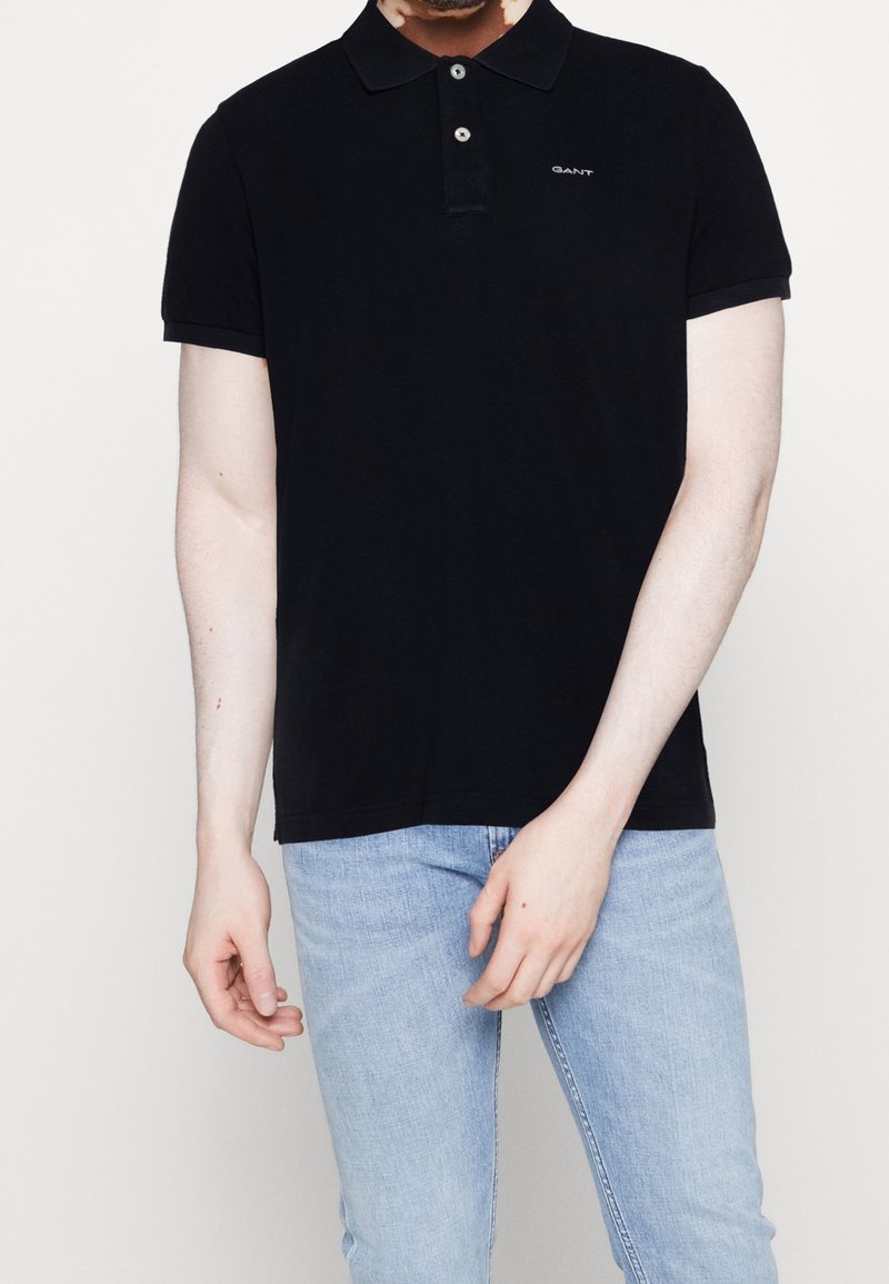 Polo noir à manches courtes avec col, comprenant une patte de boutonnage à trois boutons et un logo discret. Porté avec un jean bleu clair.