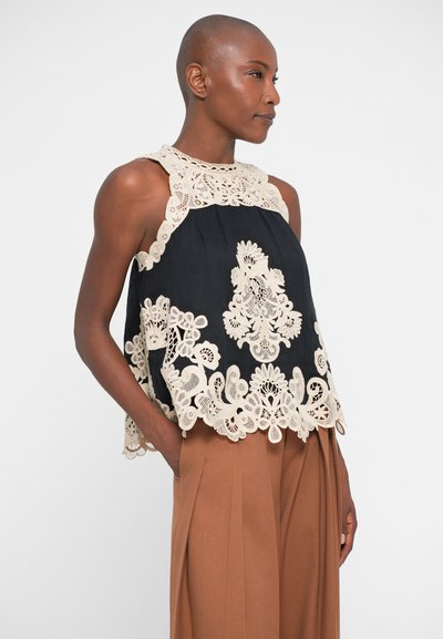 Top preto com padrão floral de renda em creme, apresentando um pescoço alto e um corte mais folgado. Combinado com calças pregueadas castanhas.