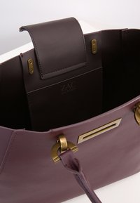 Borsa in pelle bordeaux con hardware dorato. L'interno presenta un logo, una chiusura magnetica e un vano spazioso. Texture liscia.