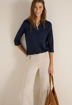 Smilende kvinde iført en marineblå top med trekvartlange ærmer og beige bukser med vide ben, der holder en brun ruskindstaske mod en ensfarvet væg.