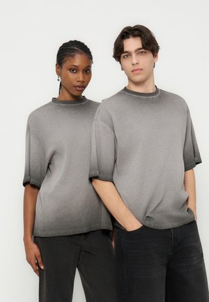 Dvaja modelky stoja vedľa seba a majú na sebe zladené sivé pletené tričká s krátkym rukávom a čierne nohavice na jednoduchom bielom pozadí.