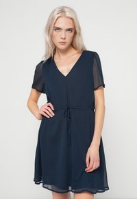 Νεαρό άτομο με ανοιχτό ξανθό μαλλί, φορώντας ένα ναυτικό μπλε φόρεμα με V ντεκολτέ και διαφανή κοντά μανίκια, με δεμένο στη μέση, που στέκεται μπροστά σε ουδέτερη πλάτη.