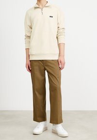 Sudadera beige con cremallera, bolsillo frontal y logo negro, combinada con pantalones marrón claro y zapatillas blancas, sobre un fondo neutro.