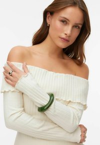 Krēmkrāsas, uzribots džemperis ar atsegtiem pleciem un volānu maliņām, rokā ar spīdīgu tumši zaļu aproci un sudraba gredzenu.