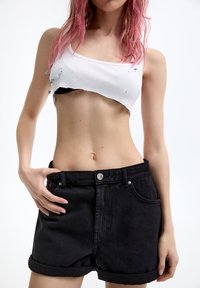 Shorts de mezclilla negra con dobladillos doblados y bolsillos frontales, combinados con una camiseta sin mangas blanca de canalé cropped que presenta acentos de imperdibles.