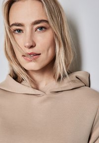 Sudadera con capucha en un color beige claro, con una textura suave y un diseño sencillo, con un escote minimalista y costuras de tela visibles.