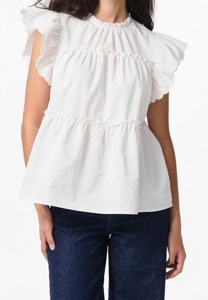 Kvinde iført en hvid ærmeløs bluse med flæser og lag, kombineret med mørkeblå jeans, står mod en ensfarvet baggrund.