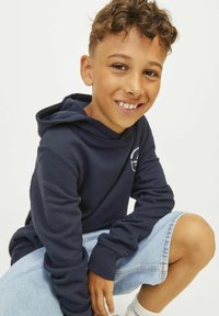 Námornícky modrá mikina s kapucňou a rebrovanými manžetami, zdobená logom, v kombinácii s svetlými džínsovými šortkami. Model sa usmieva a je v podsede.