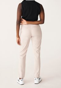 Osoba stojící zády k fotoaparátu, oblečená v světle béžových kalhotách, černém topu bez rukávů a bílých teniskách Nike Air na jednoduchém pozadí.