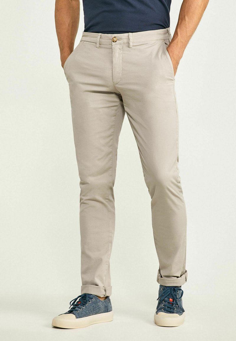 Beige cotton pants with a slim fit, featuring a front button closure, side pockets, and rolled hems, paired with blue sneakers.