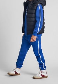 Tuta blu con strisce laterali bianche, gilet imbottito nero e scarpe da ginnastica bianche con accenti rossi. Tessuto morbido e design aderente.