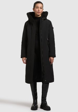 Sort lang frakk med lynlås og hætte i pels. Har sidelommer og glat, mat materiale. Båret over et sort outfit.