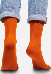 Pomarańczowe skarpetki z prążkowanego bawełnianego materiału, odsłaniające okolice kostki, noszone z lekko podwiniętymi niebieskimi dżinsami, ukazujące teksturę i kontrast kolorystyczny.